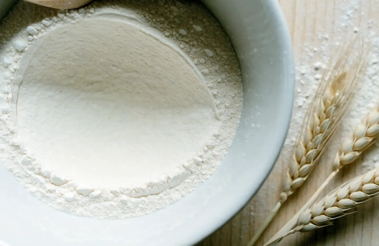 Ceramic bowl of fine white wheat flour on a wooden counter, with wheat ears beside it