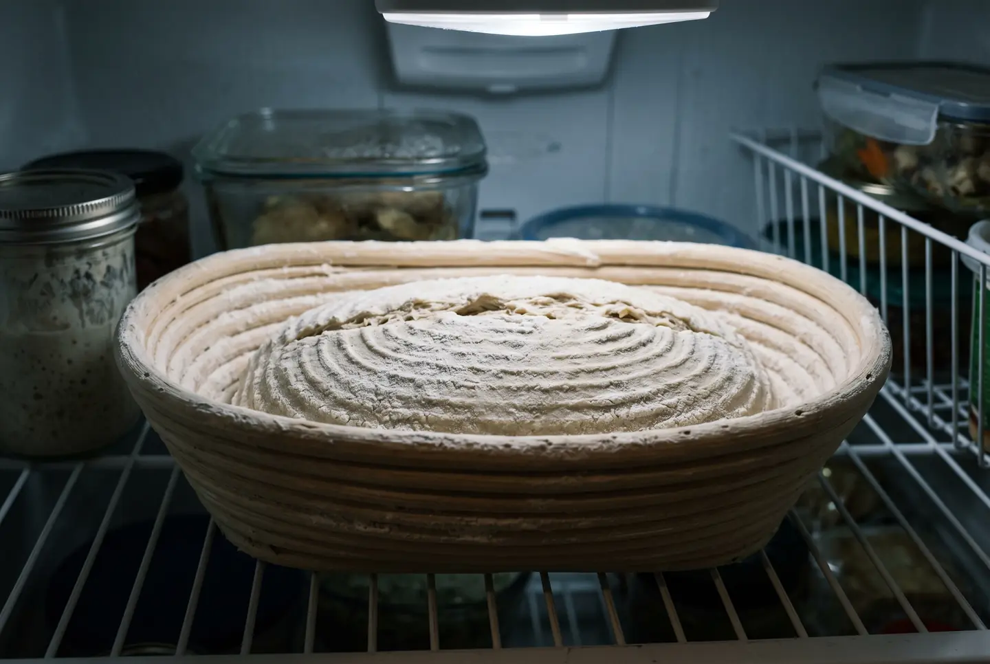 A shaped sourdough loaf resting seam-side up in a floured oval banneton on a refrigerator shelf, the surface firm and cold, ready to be scored straight from the fridge