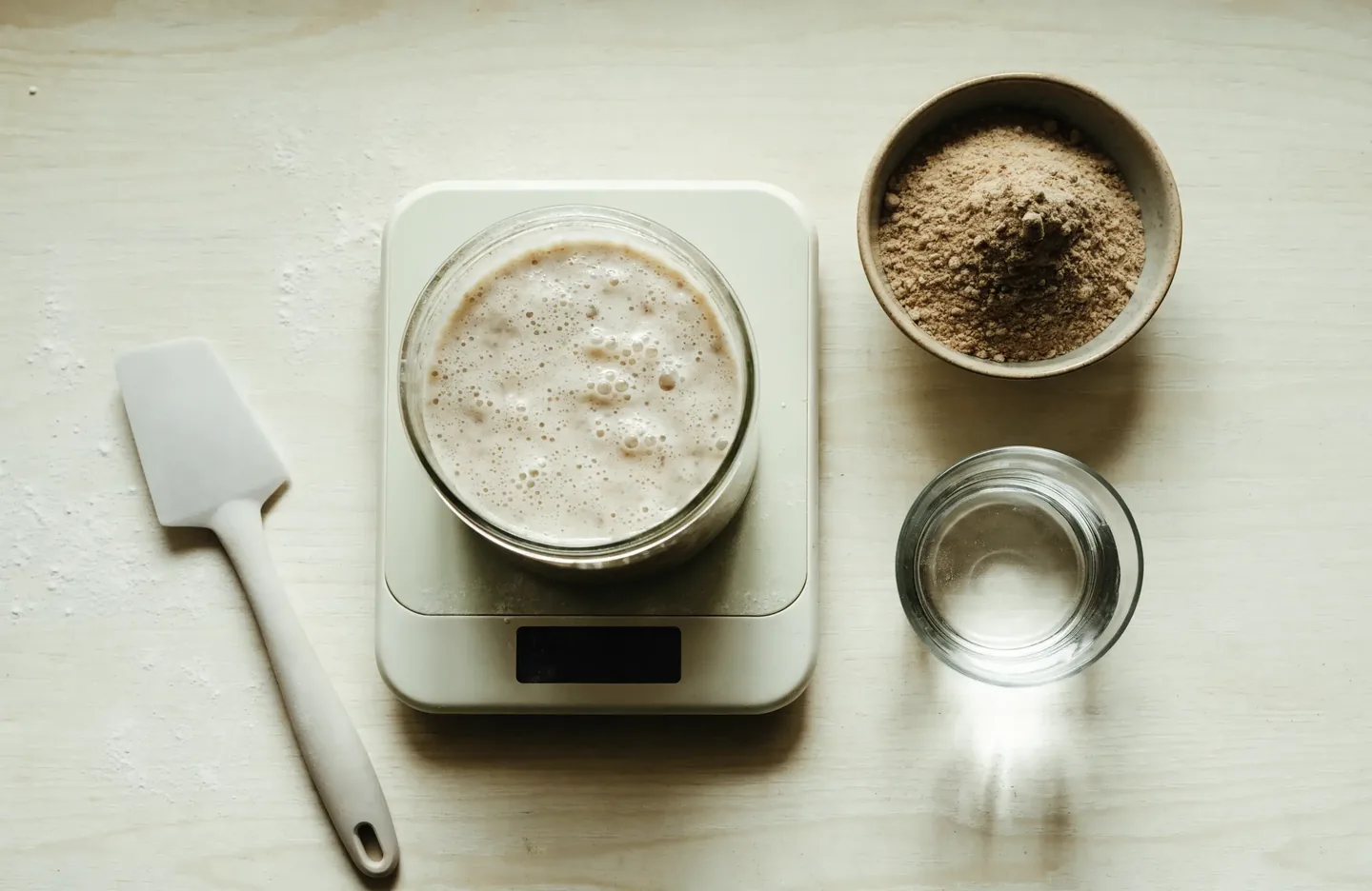 A kitchen scale holding a jar of starter beside a bowl of wholemeal flour and a glass of water, ready for a feeding measured by weight