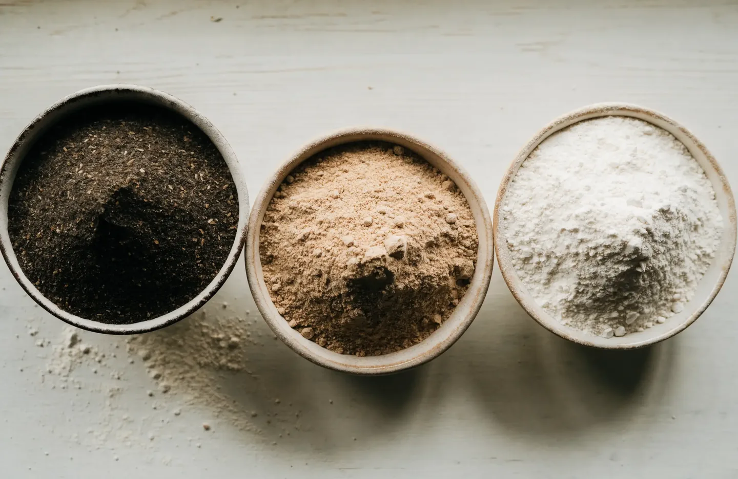 Three bowls side by side holding wholegrain rye, wholemeal wheat and white bread flour, showing the difference in colour and grain